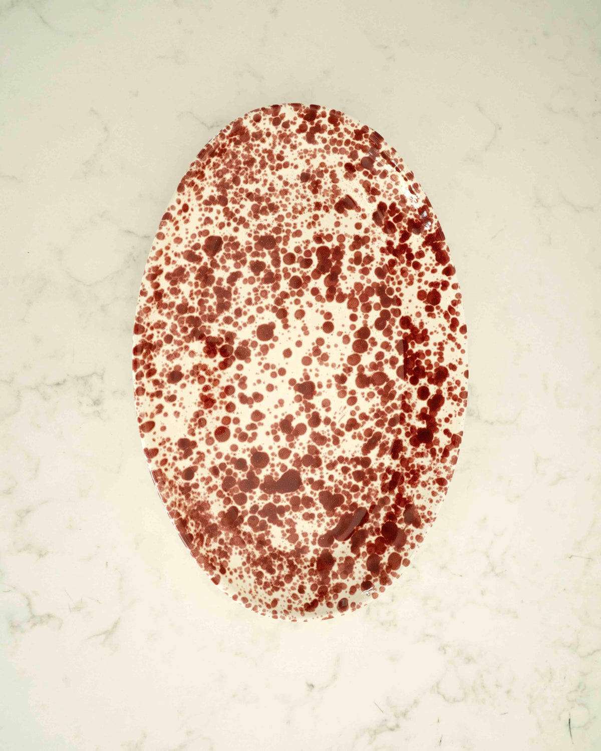 An oval white plate or tray covered in an all-over dark red speckled pattern, photographed from above on a light marble-like surface.