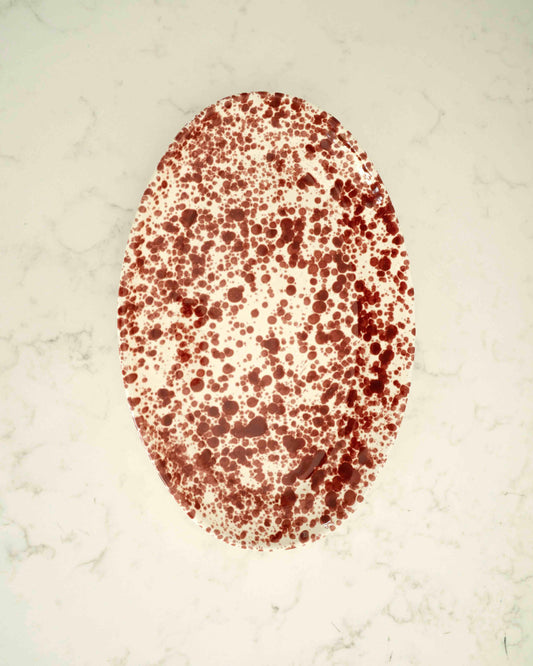 An oval white plate or tray covered in an all-over dark red speckled pattern, photographed from above on a light marble-like surface.