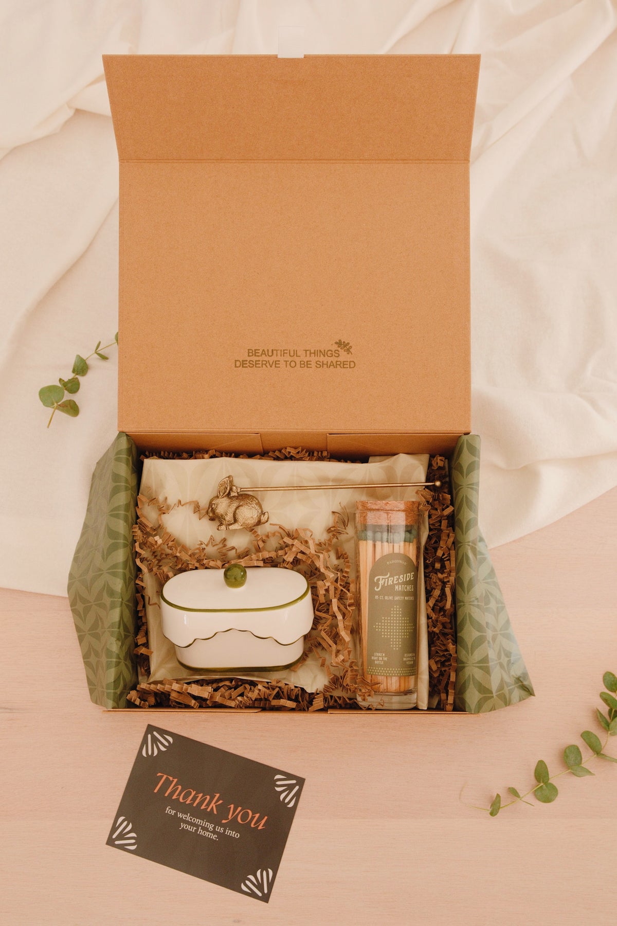 An open kraft gift box on a light wood surface with white fabric in the background, containing a small white ceramic container with a green lid, a clear pouch with cotton and botanical elements, and a tall amber bottle. A black card reading 'Thank you' is placed in front, with green leaves around the box.