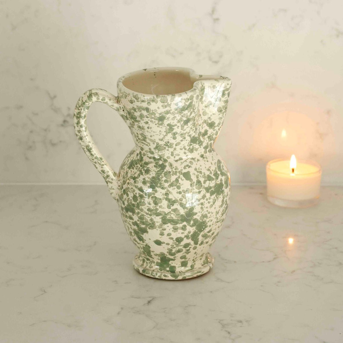 A splatter ceramic pitcher with a green and white marbled pattern, set on a white marble surface next to a lit candle against a matching marble wall.